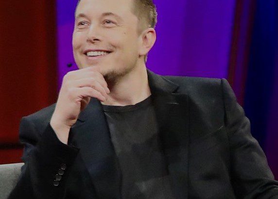 Elon Musk sorri, sentado, com a mão no queixo. Fundo roxo.