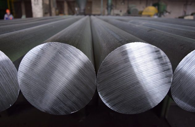 Barras de metal cilíndricas em uma fábrica, com as extremidades circulares de superfície usinada em foco. Pessoa ao fundo.