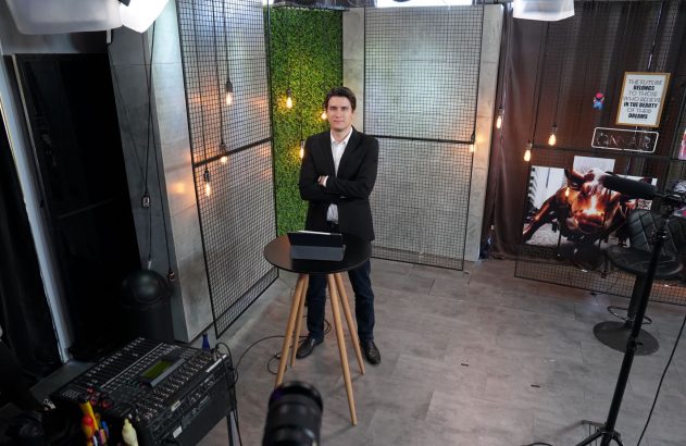 Homem de terno e braços cruzados em estúdio. Uma mesa com tablet à sua frente.