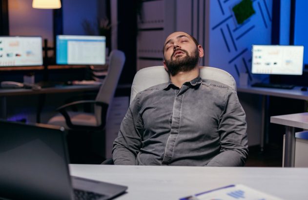 Homem cochilando em cadeira de escritório, com monitores ligados ao fundo.