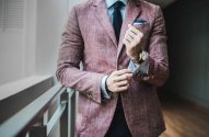 Homem ajustando o punho da camisa. Veste blazer rosado, camisa azul, gravata escura e relógio.