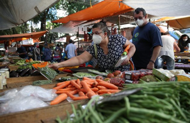 Em uma feira, uma mulher de máscara escolhe vegetais como cenouras e vagens. Um homem de máscara a acompanha.