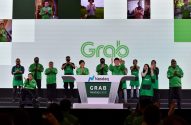 Pessoas com roupas Grab aplaudem em palco diante de pódio Nasdaq. Telas mostram outras pessoas aplaudindo. Evento de listagem.