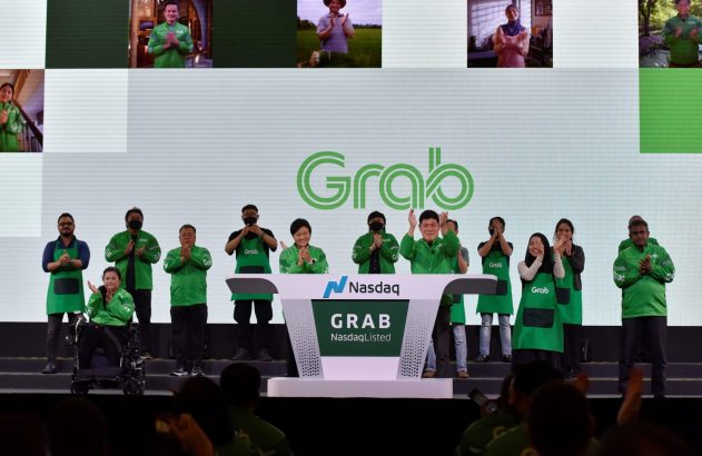 Pessoas com roupas Grab aplaudem em palco diante de pódio Nasdaq. Telas mostram outras pessoas aplaudindo. Evento de listagem.