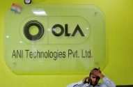 Placa de vidro da OLA/ANI Technologies em parede verde-limão, homem fala ao telefone com a mão na cabeça.