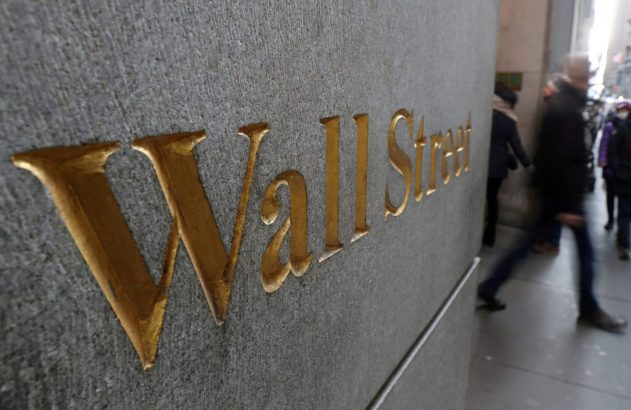Placa de pedra com o nome "Wall Street" em letras douradas e pedestres desfocados passando.