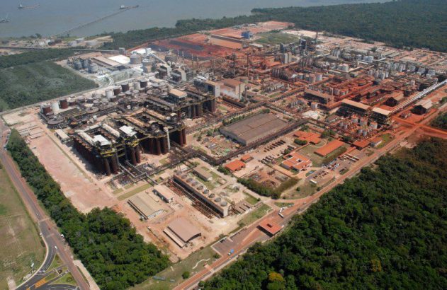 Complexo industrial gigante em operação, com porto fluvial, cercado por vasta floresta e grandes depósitos de minério.