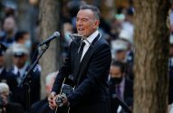 Bruce Springsteen, de terno e gravata, canta e toca violão e gaita em performance ao ar livre.
