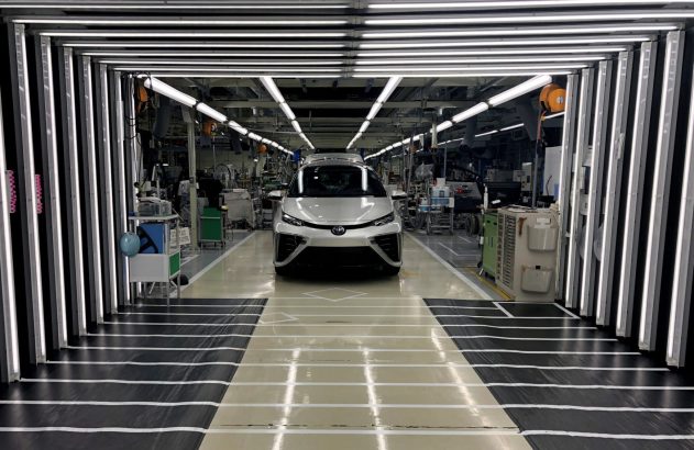 Um carro prateado é inspecionado sob luzes brilhantes em uma linha de produção automotiva.