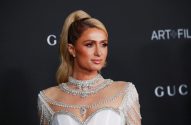 Paris Hilton, rabo de cavalo loiro, em vestido branco com joias e pérolas. Fundo escuro de evento.