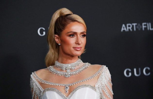 Paris Hilton, rabo de cavalo loiro, em vestido branco com joias e pérolas. Fundo escuro de evento.