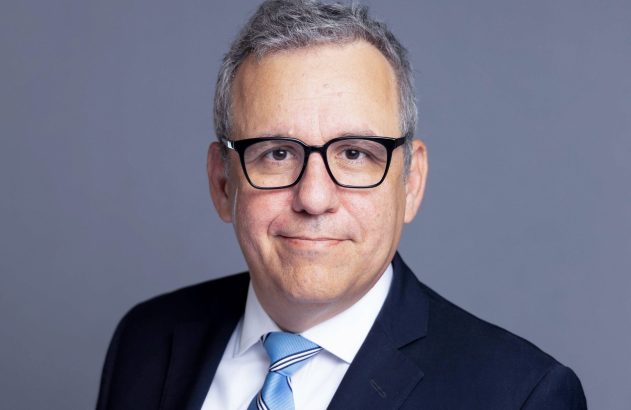 Homem de óculos, cabelos grisalhos, terno e gravata azul, sorrindo em um retrato profissional.