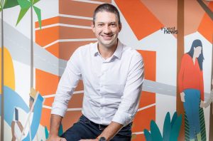 Homem sorridente de camisa branca e jeans posa contra mural colorido com logo 'invest news'.