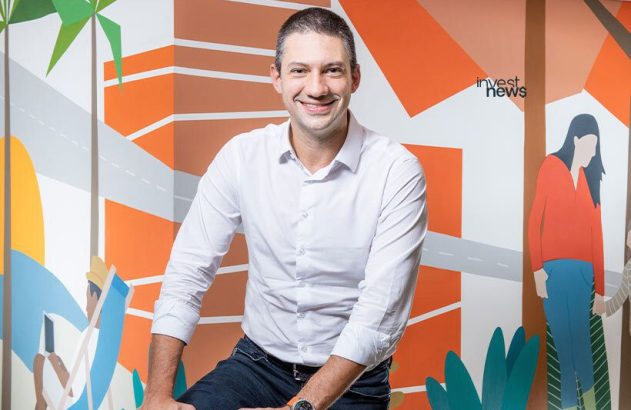 Homem sorridente de camisa branca e jeans posa contra mural colorido com logo 'invest news'.