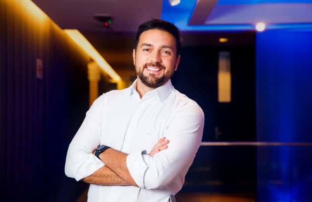 Homem sorrindo com barba, camisa branca, braços cruzados, em fundo de corredor iluminado em tons quentes e azuis.