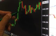 A imagem mostra uma mão apontando uma caneta em frente a um monitor, que exibe um gráfico de crescimentos e quedas como exemplo das small caps.