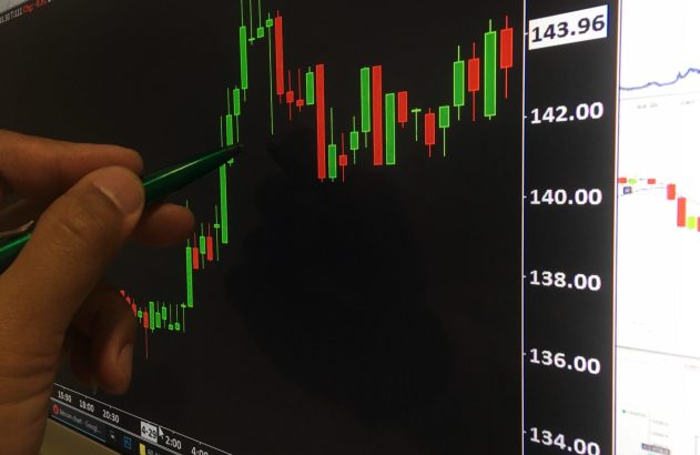 A imagem mostra uma mão apontando uma caneta em frente a um monitor, que exibe um gráfico de crescimentos e quedas como exemplo das small caps.