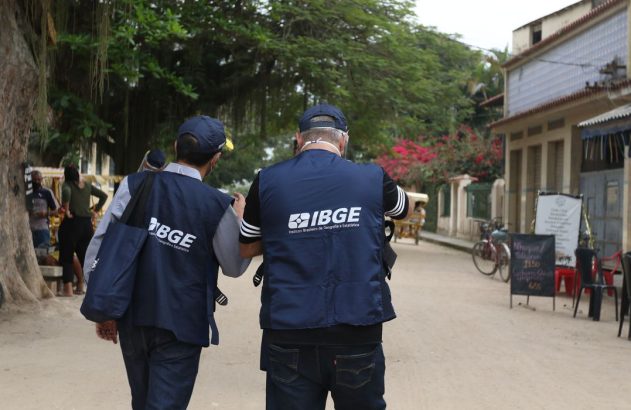 Dois recenseadores do IBGE, de coletes, caminham de costas por uma rua para coletar dados.