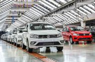 Carros Volkswagen brancos e um vermelho em fila, em linha de montagem de fábrica iluminada.