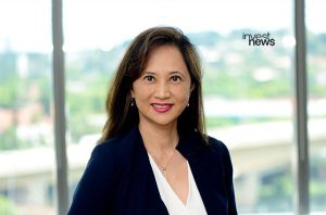 Retrato de mulher profissional sorrindo, em blazer escuro, com fundo de janelas e prédio.