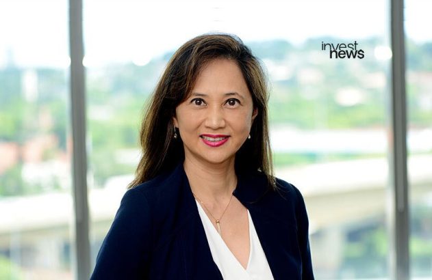 Retrato de mulher profissional sorrindo, em blazer escuro, com fundo de janelas e prédio.