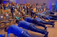 Grupo de pessoas de camisa azul fazendo flexões em um evento, com a plateia assistindo.
