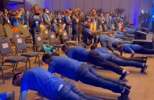 Grupo de pessoas de camisa azul fazendo flexões em um evento, com a plateia assistindo.