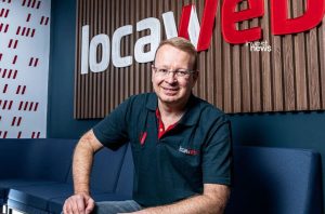 Homem sorridente sentado, com camisa Locaweb, à frente do logo da empresa.