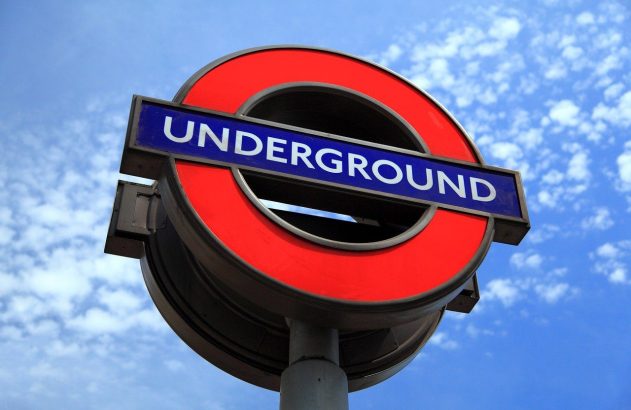 Sinal do metrô de Londres, "UNDERGROUND", em vermelho e azul, contra um céu azul com nuvens.