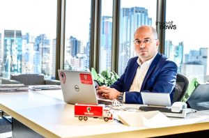 Homem em terno azul sentado em escritório, com laptop, celular e miniatura de caminhão 'Vamos' na mesa.