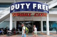 Dois homens andam na calçada em frente à loja "Duty Free Sport City", com motos estacionadas.