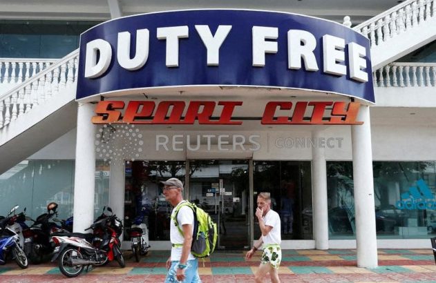 Dois homens andam na calçada em frente à loja "Duty Free Sport City", com motos estacionadas.