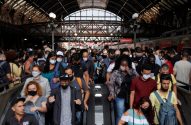 Pessoas usando máscaras descem escadas rolantes em estação de trem lotada.
