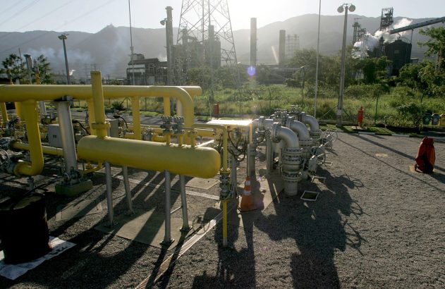 Planta industrial de gás com tubulações e chaminés ativas, montanhas ao fundo, sob luz solar.