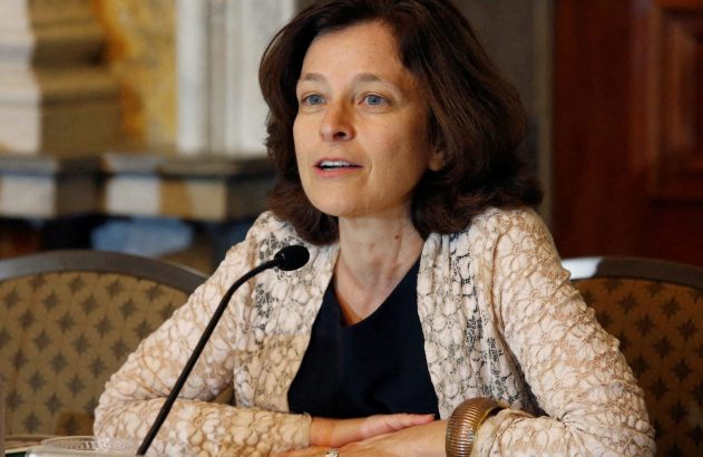 Mulher de cabelos castanhos e olhos azuis, em renda clara, falando em microfone à mesa.