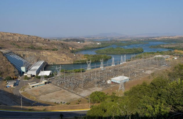 Vista aérea de usina hidrelétrica com barragem, casa de força e complexa subestação elétrica junto a rio em paisagem árida.