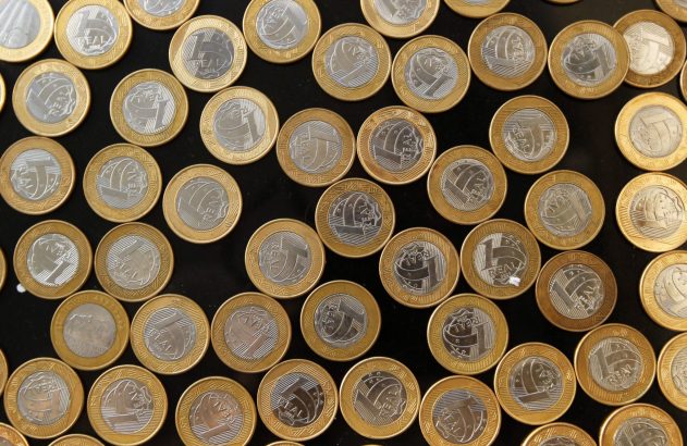 Muitas moedas brasileiras de 1 Real espalhadas sobre uma superfície escura.