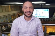 Homem sorrindo com barba e camisa xadrez em ambiente profissional, com telas ao fundo.