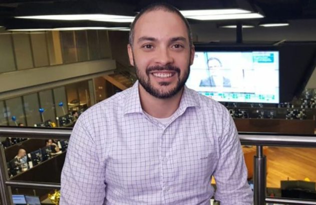 Homem sorrindo com barba e camisa xadrez em ambiente profissional, com telas ao fundo.