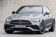 Mercedes-Benz C-Class cinza, visão frontal-esquerda. Carro estacionado com grade e faróis acesos.