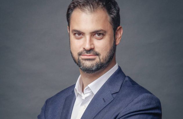 Homem barbudo, de terno azul e camisa branca, olhando para a câmera em fundo cinza.