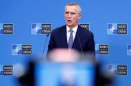 Homem de terno e óculos fala em pódio com microfones. Ao fundo, parede azul com logos NATO/OTAN. Conferência de imprensa.