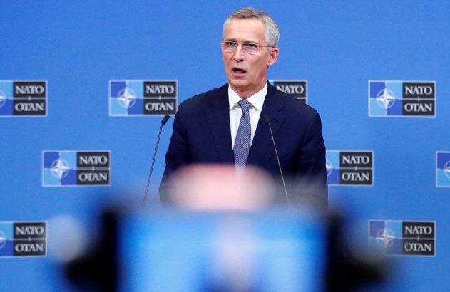 Homem de terno e óculos fala em pódio com microfones. Ao fundo, parede azul com logos NATO/OTAN. Conferência de imprensa.