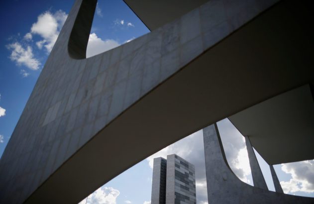 Formas arquitetônicas de concreto em primeiro plano, enquadrando prédio alto e céu nublado. Perspectiva inferior.