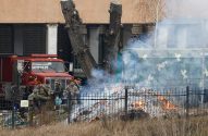 Militares se reúnem perto de uma fogueira com fumaça densa, atrás de uma cerca. Caminhão de bombeiros vermelho e veículo camuflado.