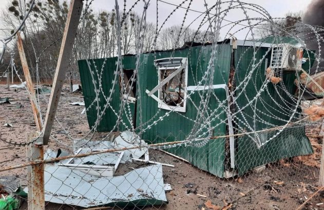 Módulo pré-fabricado verde destruído com arame farpado emaranhado em primeiro plano e detritos no chão.