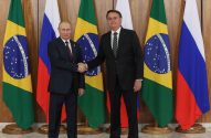 Putin e Bolsonaro apertam as mãos diante das bandeiras do Brasil e Rússia em encontro diplomático.