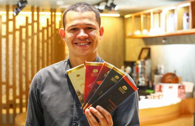 Homem sorridente de camisa escura segura várias barras de chocolate com rótulos coloridos. Fundo de loja com estantes.