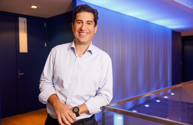 Homem sorridente de camisa azul e calça escura, encostado em corrimão, em ambiente interno com luz azul.