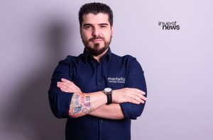 Homem barbudo de camisa azul escura com logo "martello educação financeira", braços cruzados e tatuagens.